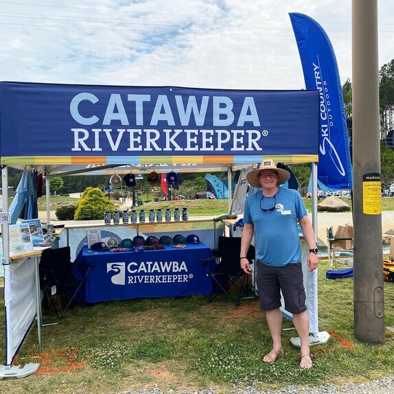 A man standing in front of a printed flat roof canopy tent at an outdoor environmental festival. 