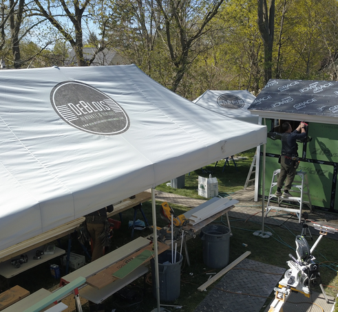 La foto mostra tre gazebo pieghevoli Mastertent con il logo DeBlois Renovate & Remodel. Un artigiano è in piedi su una scaletta ed esegue dei lavori su una casa.