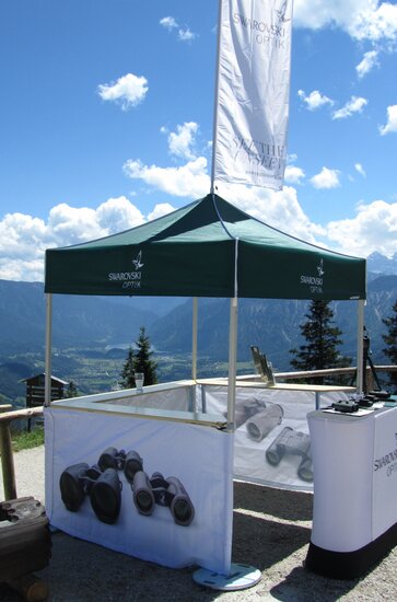 A green 5x5 canopy tent with custom printing, sidewalls, counters and a peak flag serves as an outdoor marketing stand. 