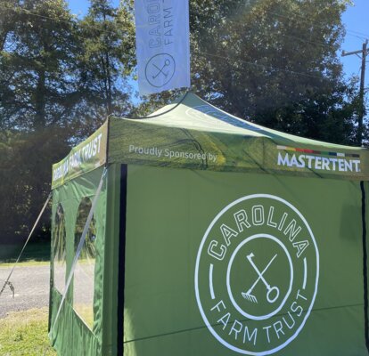 A green, 10x10ft Mastertent Canopy Tent printed with Carolina Farm Trust branding. Equipped with tent sidewalls and a peak flag.