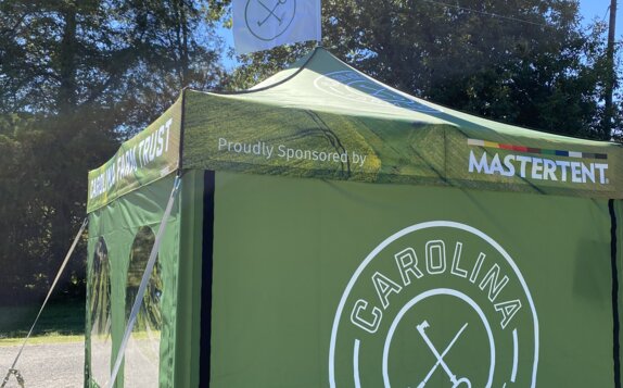 A green, 10x10ft Mastertent Canopy Tent printed with Carolina Farm Trust branding. Equipped with tent sidewalls and a peak flag.