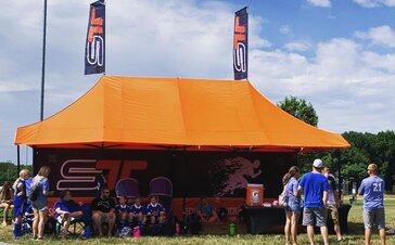 An orange Mastertent canopy tent with awnings, flags, and printed back wall set up at a sporting event for team concessions and shelter.