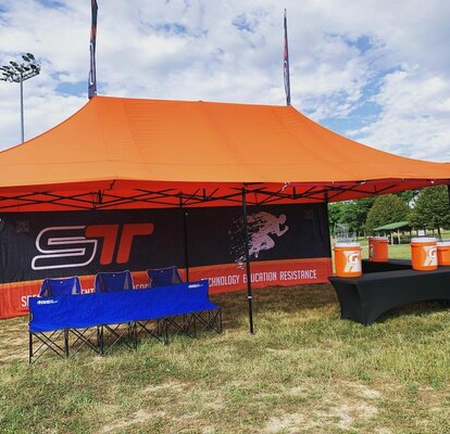 An orange Mastertent canopy tent with awnings, flags, and printed back wall set up at a sporting event for team concessions and shelter.