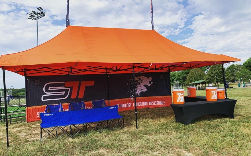 An orange Mastertent canopy tent with awnings, flags, and printed back wall set up at a sporting event for team concessions and shelter.