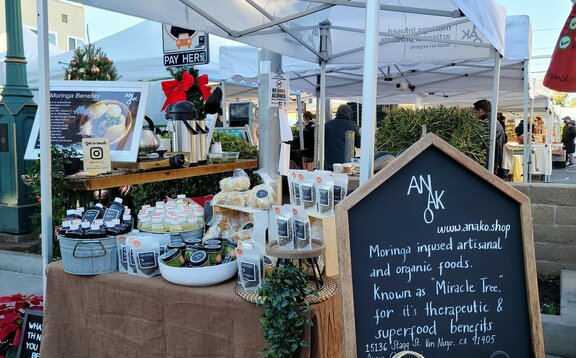 A white 10x5ft Mastertent canopy tent set up at a market to sell Anako moringa infused products at a farmer's market.