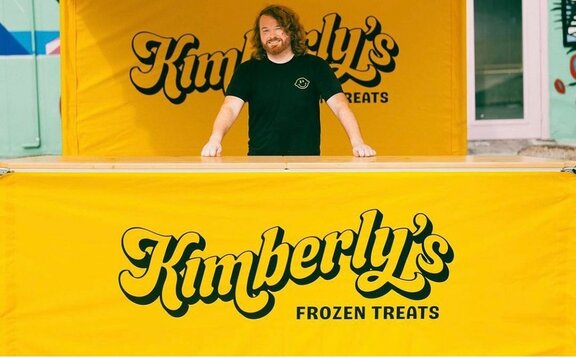 A man standing behind a counter for a yellow, 10x10ft lemonade tent with a scalloped roof, custom printing, and yellow sidewalls.