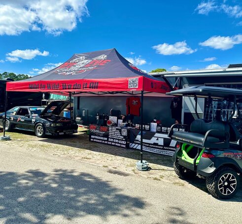 A 26x13 tent with custom printing, base weights and sidewalls serves as a work tent as it shades a car engine and mobile equipment for repairs. 