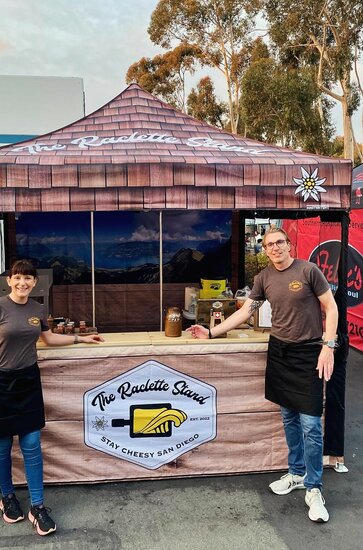 Two people stand in front of a Mastertent 10x10 custom printed canopy tent selling food at a market. 