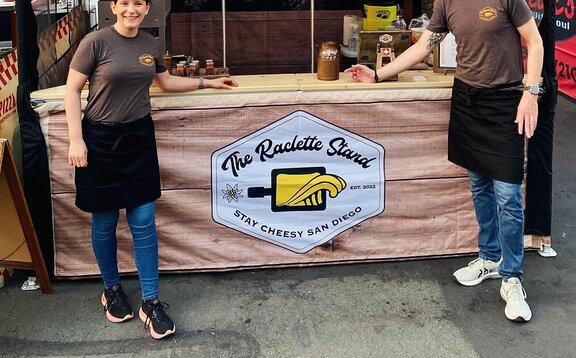 Two people stand in front of a Mastertent 10x10 custom printed canopy tent selling food at a market. 