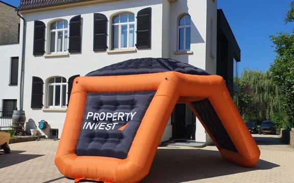 Branded black and orange tripod advertising inflatable in front of scenic house. 