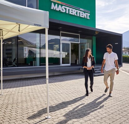 Man and woman talking and walking from modern, green building toward a white canopy tent on a sunny day