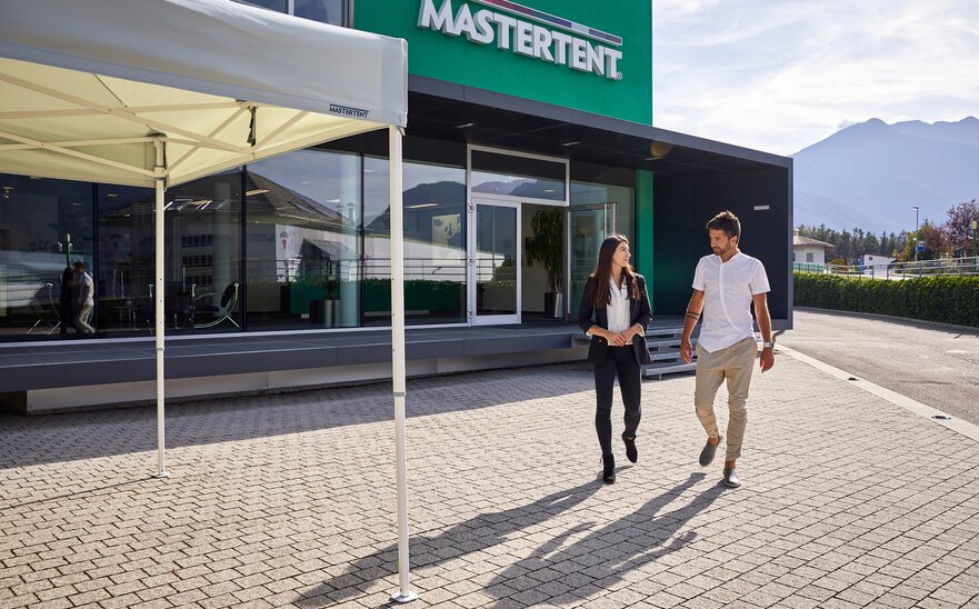 Man and woman talking and walking from modern, green building toward a white canopy tent on a sunny day