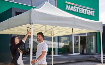 Man and woman talking and looking at a white canopy tent on a sunny day with a modern, green building in background