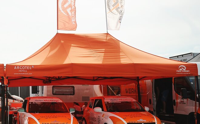 Large orange Mastertent tent with flags over two orange and white Suzuki racing cars 