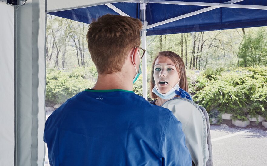 The employee inside the tent performs a smear test through the gloves integrated in the tent. The woman is in front of the test cabin.