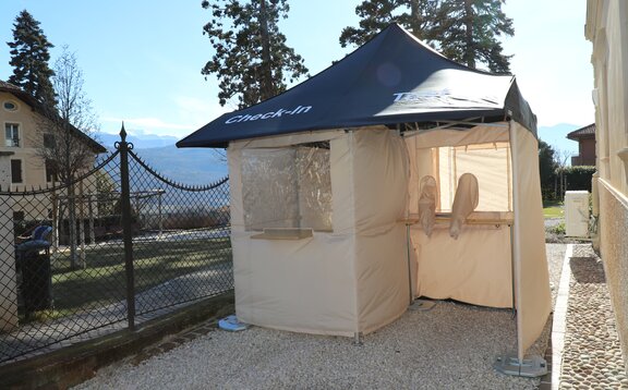 Die Covid-19 Serien-Test-Station steht vor der Apotheke Kaltern in der Goldgasse. Das Dach ist blau, die Seitenwände grau und am Himmel strahlt die Sonne.