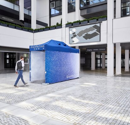 Vor einem Gebäude steht ein blauer Desinfektionstunnel. Die Person ist gerade dabei dort hindurch zugehen, damit er von Kopf bis Fuß desinfiziert wird und somit geschützt ist.