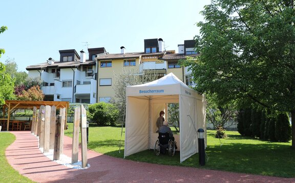 Ein mobiler Besucherraum für das Altersheim. Der ältere Herr sitzt auf einer Seite des Raums und dahinter steht die Frau. Beide sind durch eine transparente Wand getrennt.