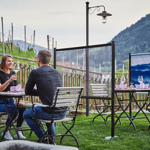 Trennwand steht zwischen den Tischen im Gastgarten. Das Paar sitzt an einem gedeckten Tisch. Dahinter sieht man noch eine bedruckte Trennwand.