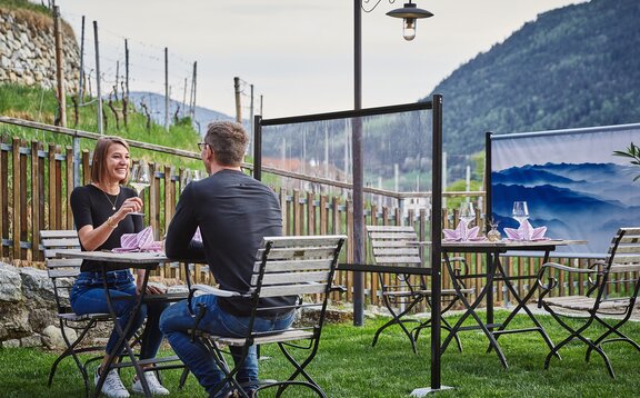 Trennwand steht zwischen den Tischen im Gastgarten. Das Paar sitzt an einem gedeckten Tisch. Dahinter sieht man noch eine bedruckte Trennwand.