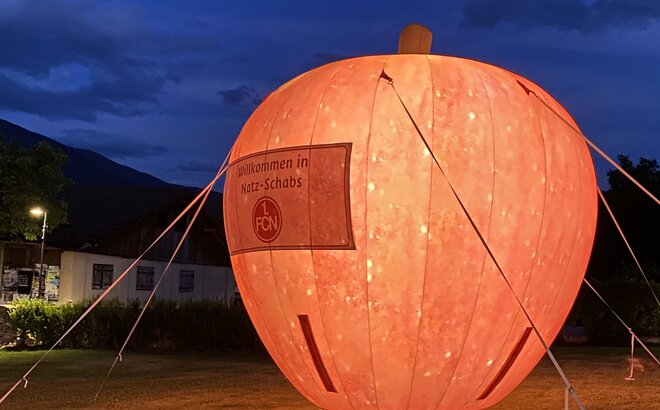 Su un prato c'è una grande mela rossa illuminata. È un mezzo pubblicitario gonfiabile.