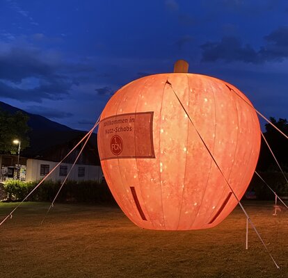 Su un prato c'è una grande mela rossa illuminata. È un mezzo pubblicitario gonfiabile.