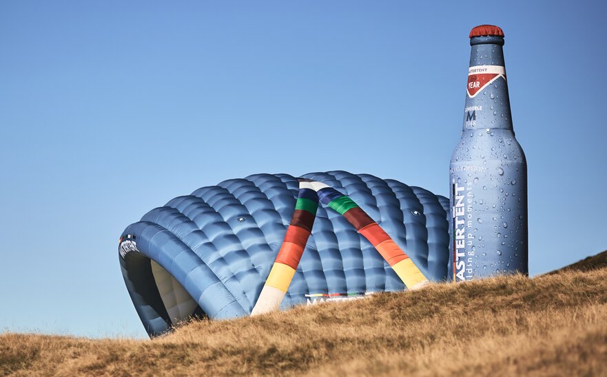 Two inflatable advertising media are located side by side on a meadow: model Kokon and model Bottle. Both in the CI colours of MASTERTENT and with a logo print.