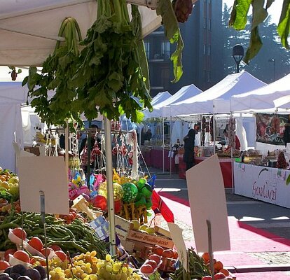 Ein Obst und Gemüse Stand steht am Bauernmarkt. Dahinter reihen sich zahlreiche weitere Marktstände. 