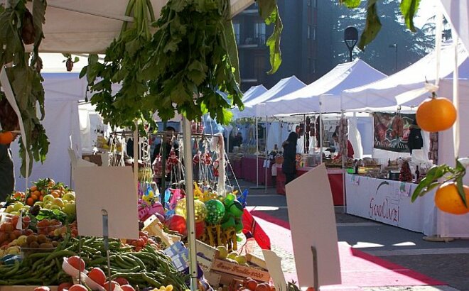 Ein Obst und Gemüse Stand steht am Bauernmarkt. Dahinter reihen sich zahlreiche weitere Marktstände. 