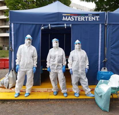 3 Sanitäter in Schutzanzügen stehen vor einem blauen Faltzelt von Mastertent. 