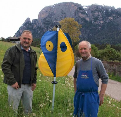 Der Kitzretter steht wie ein Regenschirm in einer Wiese. Er hat blaue und gelbe Fähnchen. Daneben stehen ein Jäger und ein Bauer. 