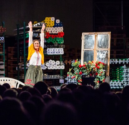 Die Schauspielerin Ingrid Maria Lechner steht auf der Bühne mit gehobenen Händen. Hinter ihr sieht man die Aluminiumstrukturen der Faltpavillons in verschiedenen Farben. 