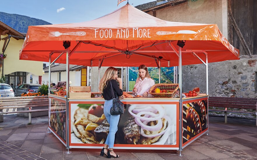 La venditrice nel chiosco esagonale offre una carota alla cliente. Il tetto del punto vendita è arancione con la dicitura "FOOD AND MORE".