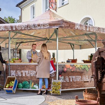 Il veditore nel punto vendita sta servendo il suo cliente. Il venditore da ad una donna un sacchetto con della frutta. Il chiosco è stampato con frutta e verdura e con la dicitura "NATURALIS".