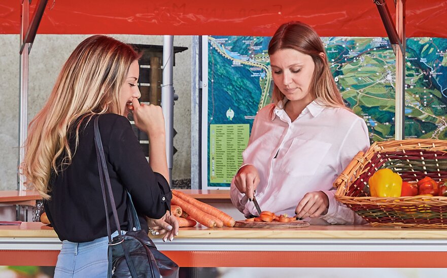 The saleswoman in the vegetable stand is cutting a carrot into thin slices. The customer in front of the pavilion is tasting a slice of carrot. 