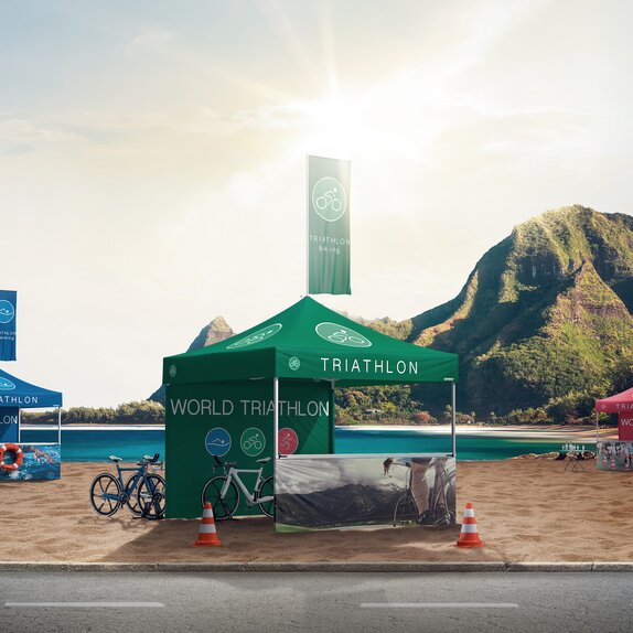 Drei Faltpavillons stehen auf dem Strand bei einem Triathlon. Die Faltpavillons sind grün, blau und rot. Alle drei haben eine Fahne und sind bedruckt. 