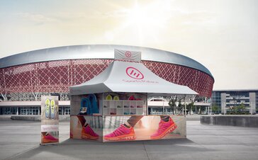 The gazebo is located in front of a stadium advertising sports shoes. The gazebo consists of a roof banner and printed sidewalls. 