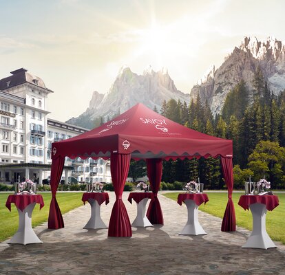 A red elegant gazebo with scalloped valance and corner curtains is located in a hotel garden. Under the gazebo are some standing tables. 