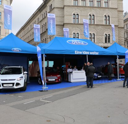 Bedruckter Werbepavillon von Ford mit vielen blauen Fahnen auf dem Dach. Unter dem Promotionzelt stehen Fahrzeuge von Ford.