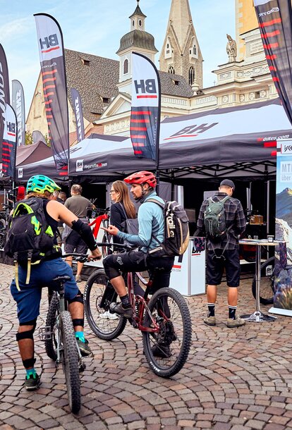 Gazebo pieghevole promozionale della ditta BH a festival delle biciclette a Bressanone. Gazebo neri e bianchi in sistema modulare personalizzati con stampa di loghi e scritte molte bandiere. Di fronte ci sono delle mountainbike. 
