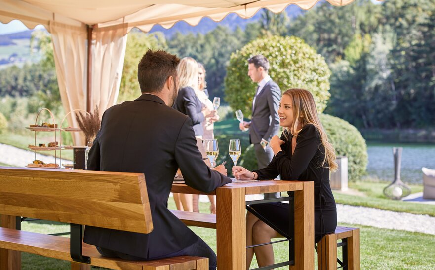 A couple is sitting under and elegant gazebo on a Lago set and is talking. Both of them are holding a glass of white wine in their hands.