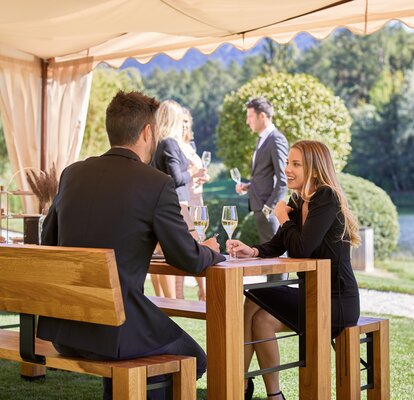 Unter einem eleganten Faltpavillon sitzt ein Paar auf einer Lago Garnitur und unterhält sich. In der Hand halten beide ein Glas Weißwein.