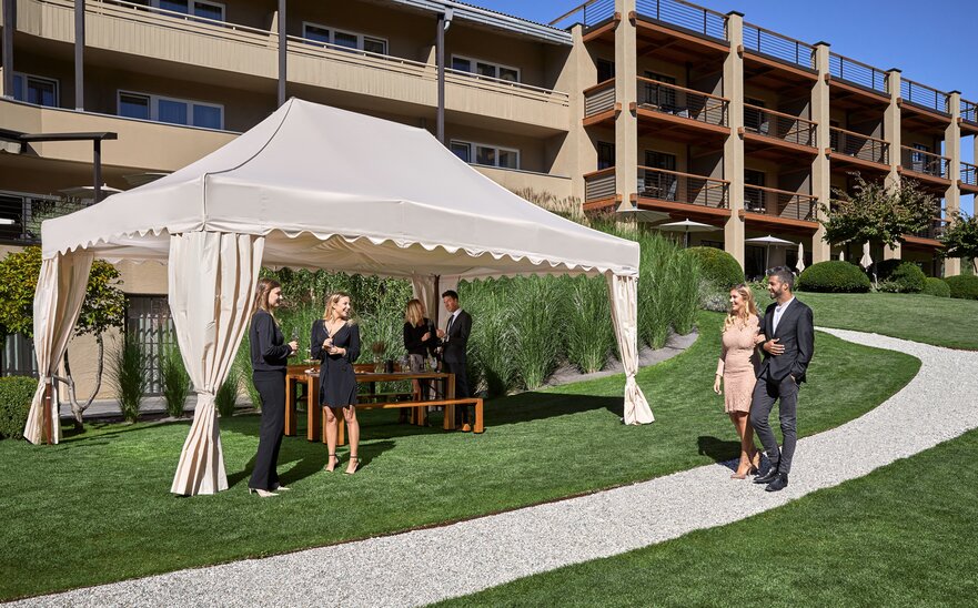 Un gazebo pieghevole elegante die MASTERTENT si trova di fronte ad un albergo. Gli ospiti sono vestiti in modo elegante e si trovano vicino al gazebo.