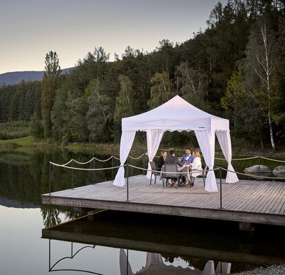 Weißer Faltpavillon steht auf dem Steg am See. Darunter dinieren 4 Personen.