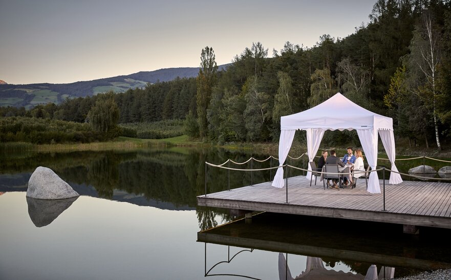 Weißer Faltpavillon steht auf dem Steg am See. Darunter dinieren 4 Personen.