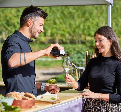 Il vignaiolo versa il vino bianco dentro il bicchiere dalla donna. Lui si trova sotto il gazebo pieghevole e lei davanti.