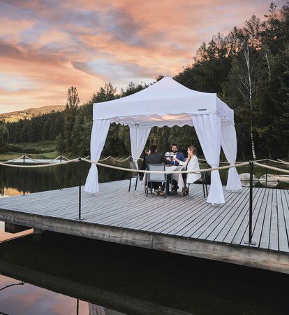 A white gazebo with corner curtains is located on the footbridge. Four people are dining underneath in the sunset. 