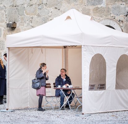 Il gazebo pieghevole per fumatori di fronte al ristorante ha 3 pareti laterali. Sutto stanno conversando una donna che fuma e un uomo che beve un caffè.