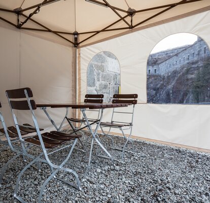 Der Faltpavillon von innen. Es steht ein Tisch mit Stühlen im Zelt. Man kann die Seitenwand mit Fenstern sehen.