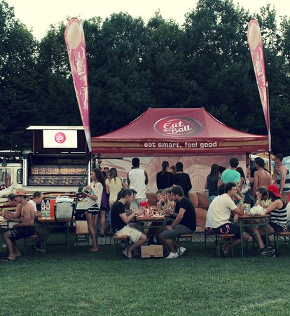 Der bordeaux rote Faltpavillon von "Eat the Ball" steht neben dem Food Truck. Davor sitzen die Menschen und essen einen Burger.